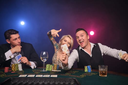 Two athletic fellows and alluring lady are playing poker at casino. Youth are making bets waiting for a big win. They are smiling and posing sitting at the table against a red and blue backlights on black smoke background. Cards, chips, money, gambling, entertainment concept.