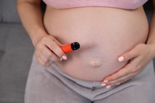 A pregnant woman smokes a vape while sitting on the couch. Close-up of the abdomen