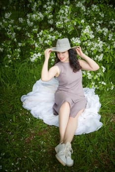 Elegant Woman Posing With Hat in Blossoming Garden. Smiling woman in a dress playfully tipping her hat among spring blossoms. The concept of fashion, self care