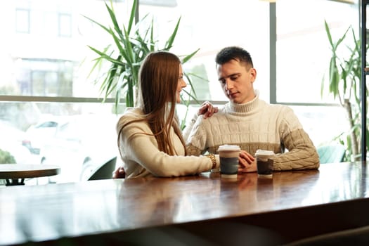 A couple sits close together at a wooden table in a brightly lit cafe, exchanging smiles over steaming cups of perhaps coffee or tea. They appear relaxed and engaged in a cheerful conversation, with natural light filtering through the space creating a warm and inviting atmosphere.