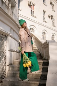 A woman wearing a green hat and green pants is holding a bouquet of yellow flowers. She is standing on a set of stairs in front of a building