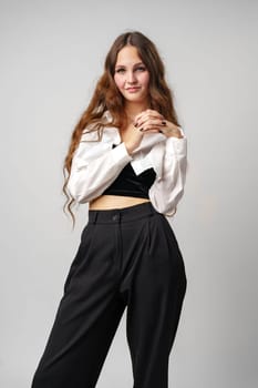 A confident young woman stands with her hands clasped together, wearing a stylish white cropped jacket paired with high-waisted black trousers. A subtle smile plays on her lips as she poses against a neutral grey backdrop, exuding a mix of casual elegance and relaxed poise.
