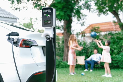 Focus electric vehicle recharge from home charging station on blur background of happy and playful family playing together. EV car using alternative and sustainable energy for better future.Synchronos