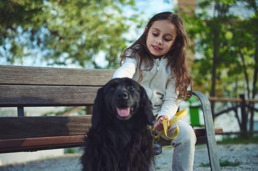 Lovely little kid girl 5-7 years old, sitting on bench and stroking her black cocker spaniel dog while walking it on leash. Playing pets. The concept of love, care and empathy for domestic animals