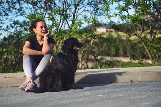 Multi ethnic beautiful thoughtful woman sitting next to her dog on the asphalt, a beautiful black cocker spaniel and looking dreamily into the distance. Pets and people. Woman walking her dog on leash