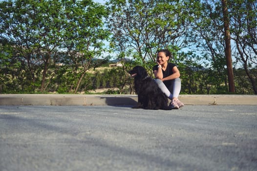 Full size portrait of woman sitting on the parapet, smiling and talking to her dog while taking her pet for a walk on the nature. People and animals concept