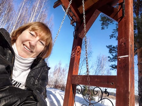 A cheerful middle aged woman in a winter coat taking selfie on the swing on nature outdoors in sunny day with blue sky