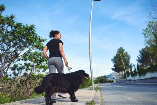 Rear view of a young active woman walking her dog on leash in the nature on a beautiful sunny summer day. Pretty female athlete running with her pet in the morning. People and domestic animals