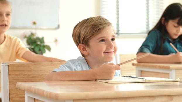 Caucasian boy raising hand for asking and answering teacher surrounded by diverse children studying at classroom. Happy student working together, voting, volunteering, calling instructor. Pedagogy.