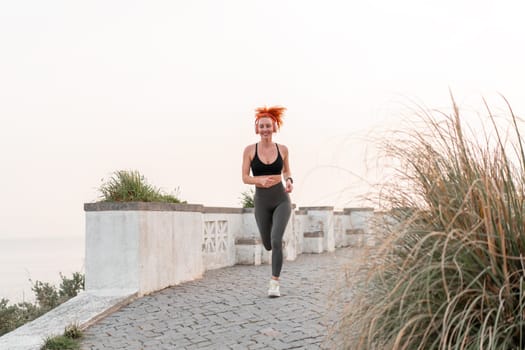 Happy redhead woman running on footpath. Full body of happy fit female runner in sportswear jogging on seaside. Woman jogger listening to music. Health and fitness conepts.