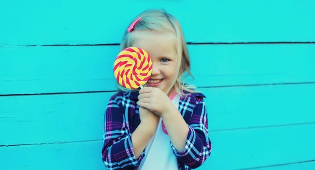 Portrait of happy cheerful smiling little girl child with sweet lollipop on stick on blue background