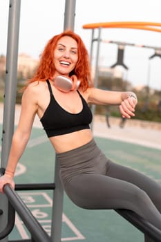 Cheerful sporty woman jogger with red hair checking time on wristwatch. Smiling fit lady runner take a break after workout running exercises in summer gym park.