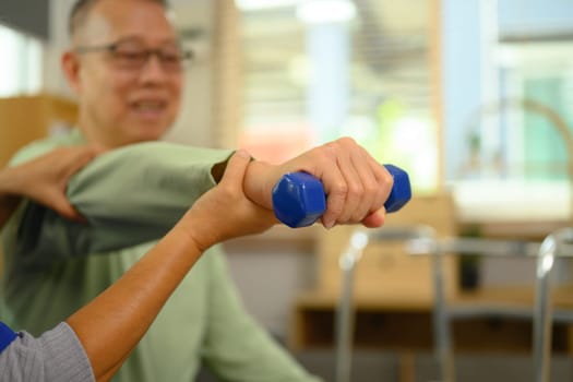 Physiotherapist helping senior man exercising with light weight dumbbell at home.