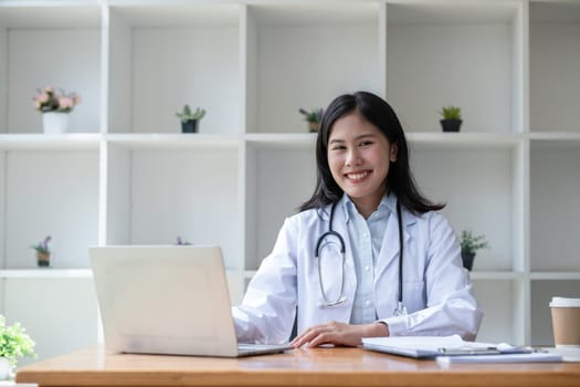 young asian lady doctor in white medical uniform with stethoscope using computer laptop talking video conference call with patient at desk in health clinic or hospital..