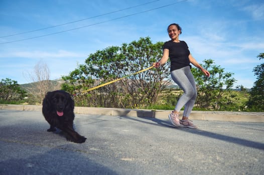 Full length authentic portrait of a cheerful happy young woman walking her dog on leash in the nature on a beautiful sunny summer day. Pretty female athlete running with her pet in the morning