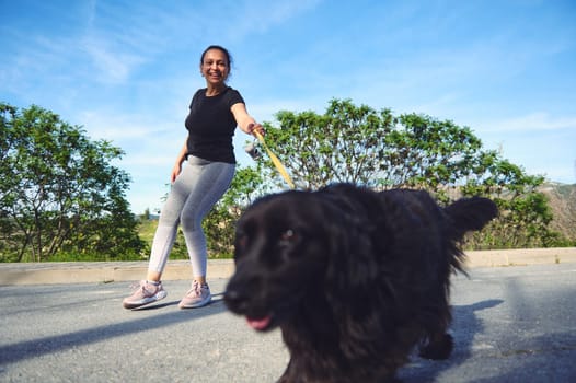 Happy woman walking her dog outdoors. Pretty female athlete dressed in gray leggings and black t-shirt, walking her cocker spaniel pet on leash. People. Animals. Nature. Playing pets concept