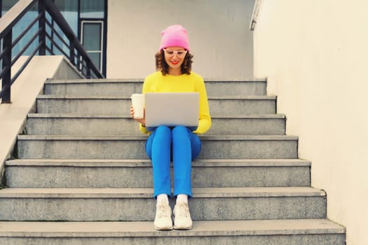 Portrait of stylish modern young woman working with laptop wearing a colorful clothes, eyeglasses in the city
