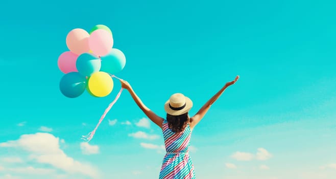 Back view of happy joyful young woman with bunch of colorful balloons in summer straw hat on blue sky clouds background