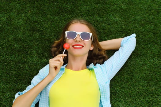 Portrait of happy teenager girl resting with lollipop lying on green grass in summer park, top view