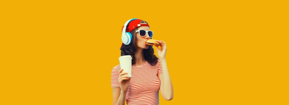 Stylish young woman listening to music in headphones eating burger fast food holding cup of coffee or juice on yellow background