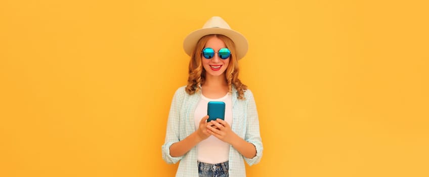 Summer portrait of happy traveler young woman with mobile phone looking at device in straw tourist hat on yellow studio background