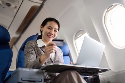 Young Asian businesswoman holding credit card in online shopping on laptop sitting near window on airplane during flight, travel and business tourism concept.