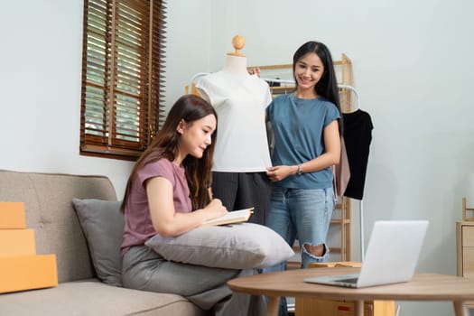 Asian woman friends couple talk to camera to sell the clothes at home. Young attractive business people female live streaming and showing clothing to customer in online.