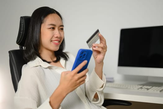 Pleased young woman with credit card in hands making internet order purchase on mobile phone.