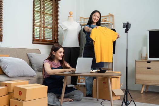 Asian woman friends couple talk to camera to sell the clothes at home. Young attractive business people female live streaming and showing clothing to customer in online.