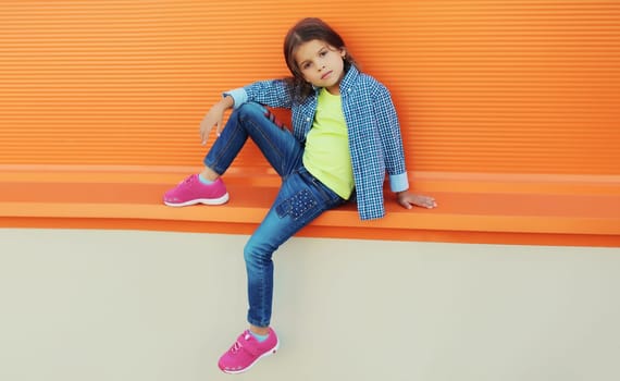 Stylish little girl child in colorful clothes posing on city street on orange wall background