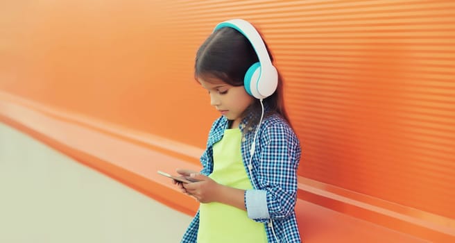 Portrait of little girl child with smartphone listening to music in blue wireless headphones, looking at device on orange background