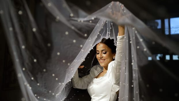 A beautiful bride holds a veil in her hands. Happy beautiful bride under a veil close-up. A bride in a white dress plays with a veil. Turkmen bride.