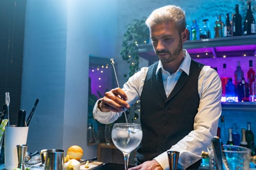 Young barman in a white shirt and black apron making cocktail at party in nightclub with colorful lights. Nightlife concept. High quality photo