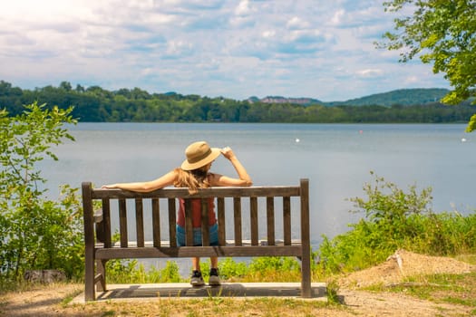 Back view of attractive young woman relaxing on a bench outdoors in front of a lake. High quality photo
