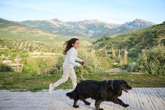 Cute school child girl in white sports clothes, running with her dog on leash in the nature, against mountains background