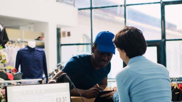 African american deliveryman carrying carton boxes, signing shipping report on tablet computer discussing orders distribution with manager. Store employee working in modern boutique