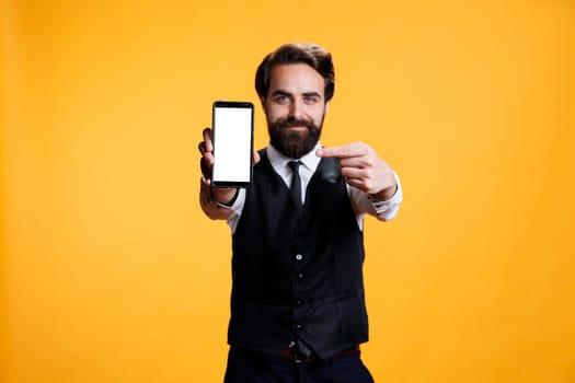 Confident waiter points to white screen on camera, presenting isolated mockup blank display on mobile phone. Young butler with suit and tie shows empty device with copyspace template.