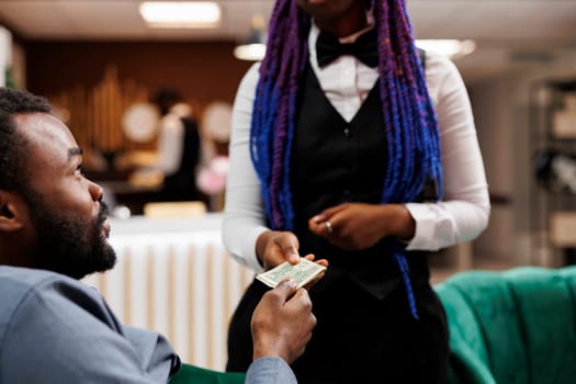 Young African American man hotel guest giving cash to waitress while relaxing in lobby, ordering food and drinks, tipping hospitality staff while resting at resort. Tipping behaviour in hospitality