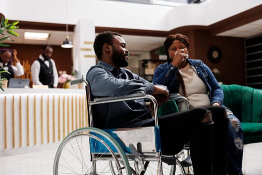 Young African American man on wheelchair sitting in hotel lobby with wife, tired unhappy woman travelling with disabled husband. Inclusive hospitality, handicap people and travel