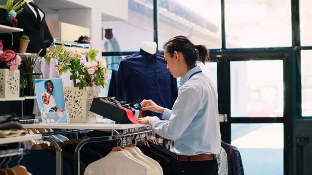 Asian store employee arranging trendy accessories, putting fashionable tie on shelf, working at modern boutique visual. Stylish man checking formal wear items in shopping mall. Fashion concept