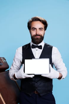 Classy hotel porter with white screen on tablet, posing on camera next to pile of baggage. Young man bellhop presenting blank gadget display, showcases professionalism of his occupation.