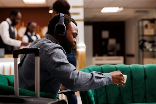 Serious African American businessman wearing headphones drinking coffee while waiting for hotel room to be ready, sitting with luggage in lobby. People and traveling, business trip concept