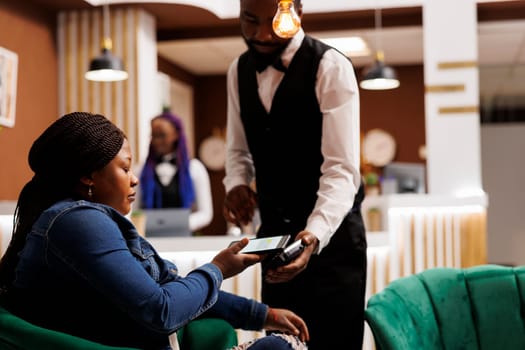 African American woman hotel guest sitting in lobby holding smartphone paying for order using nfc technology. Waiter holding POS terminal for contactless payment serving female traveler
