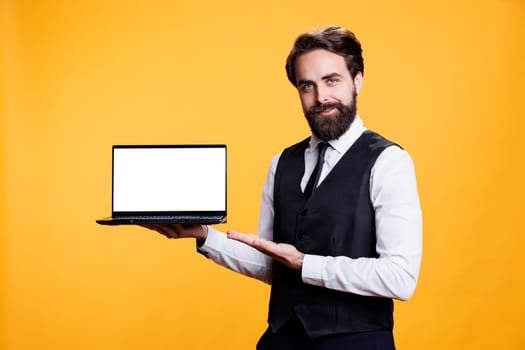 Professional man holds laptop with white display in studio, presenting isolated copyspace template on camera. Young adult employee using pc with blank empty mockup screen, yellow background.