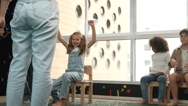 Diverse children playing musical chairs games while young smart beautiful teacher turn off music. Multicultural students running and sitting on chair in creative activity. Music lesson. Erudition.