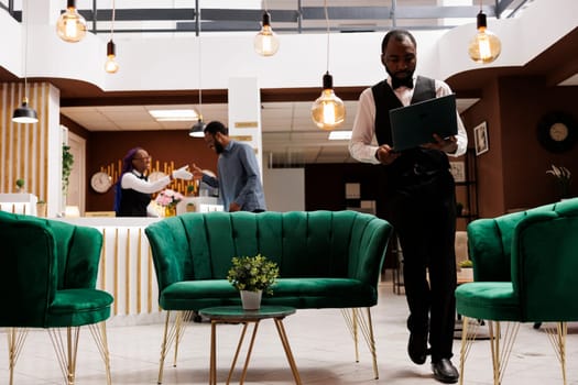 Young African American man hotel employee wearing standing uniform with laptop in lobby, using computer software program to make reservations. CRM in the hospitality industry