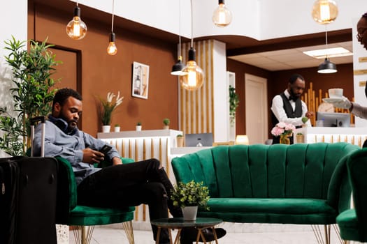 Black guy traveler sitting in hotel lobby with smartphone making mobile check-in. Young African American man tourist with luggage using phone texting sms message, waiting for room