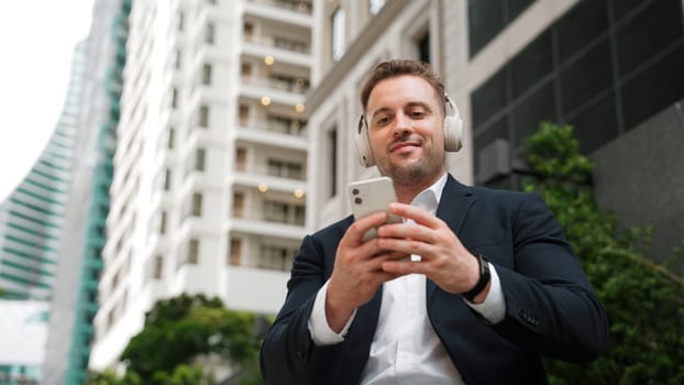 Low angle camera of project manager listening relaxing music while move to song and clap hands. Happy caucasian businessman enjoy listening music while sitting at bench at green urban city. Urbane.