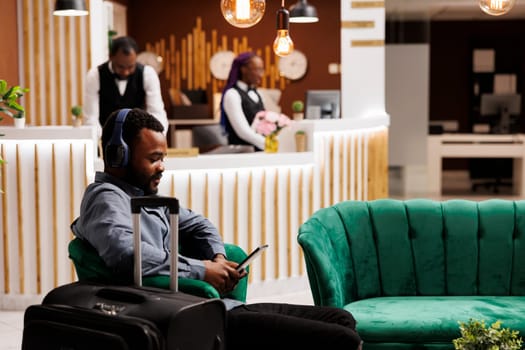 Smiling young African American man traveler waiting for room in hotel. Black guy in headphones using digital tablet while sitting in lobby with luggage, watching movie, wait for check-in process