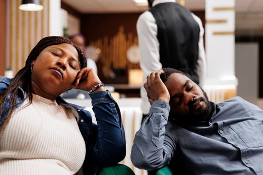 Young tired African American couple napping in hotel lobby, arriving too early at resort, resting after long flight. Exhausted tourists sleeping while traveling. People and jet lag concept
